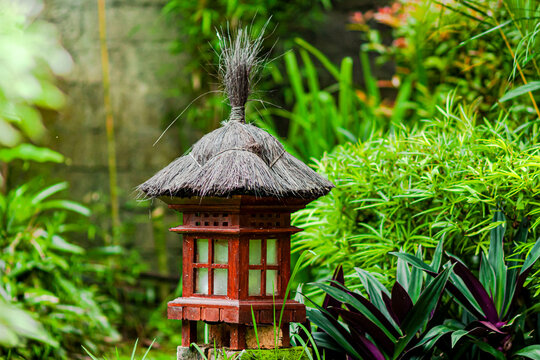 A small red wooden lantern with a thatched roof stands in a lush green garden. The intricate design and natural surroundings create a peaceful, traditional, and tropical atmosphere.