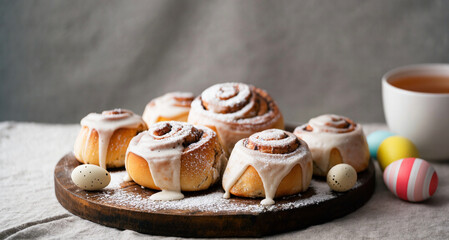 “A cozy, freshly baked batch of Easter cinnamon rolls, topped with a sweet glaze and sprinkled with powdered sugar. The rolls are placed on a wooden board next to a cup of tea and small decorative egg