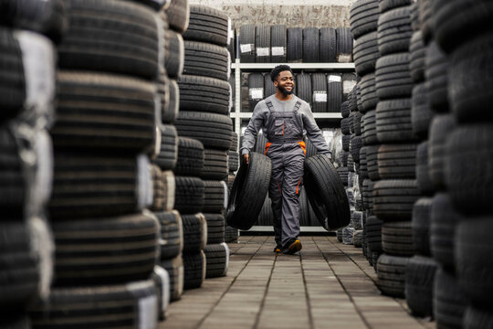 Hardworking diverse auto mechanic carrying car tires at car service surrounded by piles of tires.