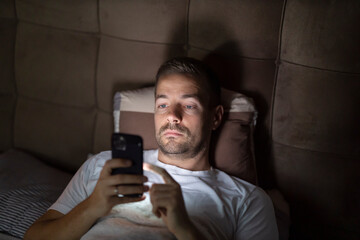 Exhausted man with insomnia lying in bed and doom scrolling on cellphone at night.
