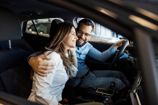 Young buyer couple sitting in classy car at car showroom and testing it while hugging.