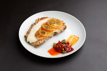 Breaded milanese cutlet topped with melted mozzarella, sliced tomato and oregano, served with roasted peppers and spicy sauce on a white plate