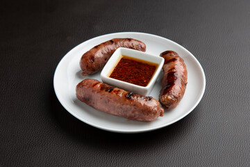 Grilled creole sausages on white plate, featuring zesty dipping sauce, epitomizing casual outdoor dining with bold, spicy flavors