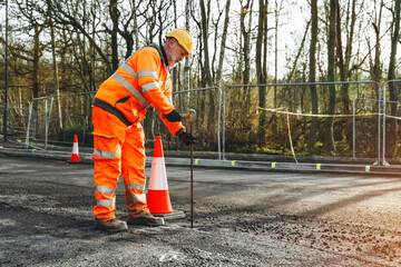 Construction worker conducting road repairs at a site wearing safety gear and using a measuring...
