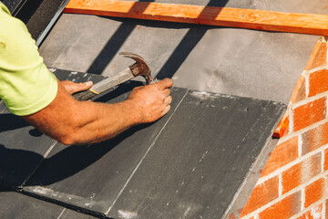 Close-up at Roof installation in progress with a worker hammering slate tiles on a sunny day at a...