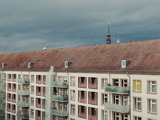 Modern residential building with balconies in Dresden, Germany. The red-tiled roof contrasts with the dark clouds, while historic church spires peek out from behind, blending old and new architecture.