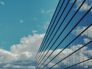 Modern glass building reflecting a bright blue sky with scattered white clouds. The sleek design and geometric perspective create a striking contrast between architecture and nature.