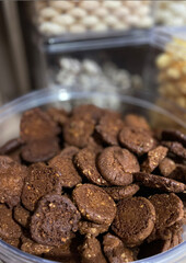 Pote transparente grande, com aproximadamente 45 cm de altura e 30 cm de diâmetro, repleto de biscoitos marrons organizados até a borda. A composição destaca o centro do pote, enquanto o fundo está le