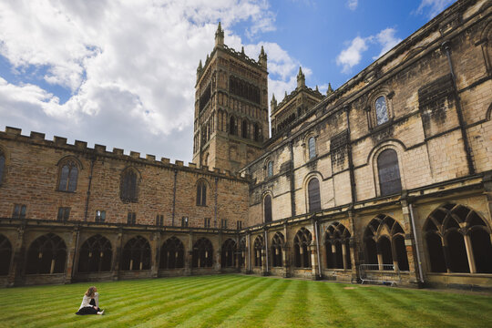 A female tourist admiring the cloisters of historic Durham Cathedral showcase its medieval arches, patterned lawn, and rich heritage under a blue sky.