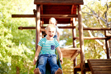 Excited, playing and portrait of children on slide in park for physical skills, confidence or fun. Exploring, nature and boy kid son playground equipment for adventure, hobby or cognitive development