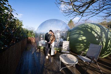 Romantic couple embracing, holding hands near transparent bubble hotel room, standing on wooden deck with garden furniture surrounding verdant landscape