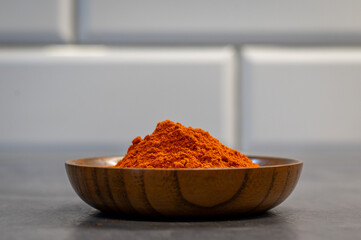A pile of vibrant red paprika powder displayed in a wooden bowl on a dark countertop. The minimalist background with white tiles emphasizes the bold color of the spice.