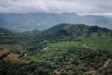 Aerial view of lush green valleys, scattered trees, winding roads, and mist-covered mountains under a cloudy sky in a tranquil rural setting.
