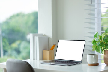 Laptop with a blank white screen sits on a white desk next to a potted green plant and a coffee mug