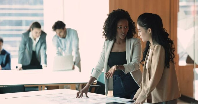 Diverse young female business colleagues reviewing and discussing sales marketing reports, standing at table with statistic paper documents. African mentor training Asian intern