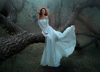 Beautiful red-haired woman in long white sitting on a tree in the forest.