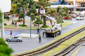 workers repairing streetlights using a crane truck near railroad tracks, convertible car parked by the sidewalk, and park area with trees and benches in the background