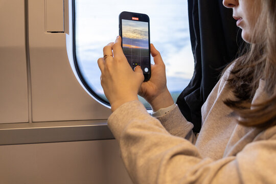 woman taking a photo of sunset through a train window using a smartphone during a train journey