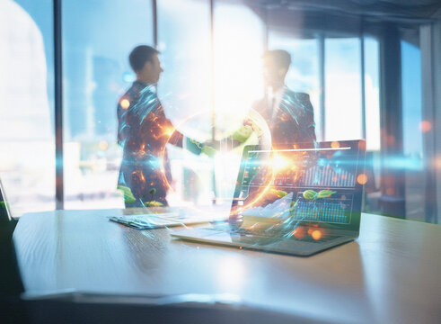 Abstract double exposure image depicts a sci-fi business deal.  Two figures shake hands, overlaid with glowing data streams on a laptop in a modern office background.