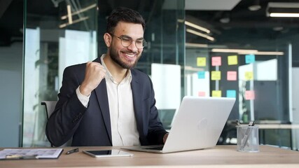 Satisfied happy businessman in a formal suit received great news on laptop while sitting at workplace in business office. Smiling glad handsome entrepreneur reads positive good message on the computer - Powered by Adobe