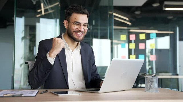 Satisfied happy businessman in a formal suit received great news on laptop while sitting at workplace in business office. Smiling glad handsome entrepreneur reads positive good message on the computer