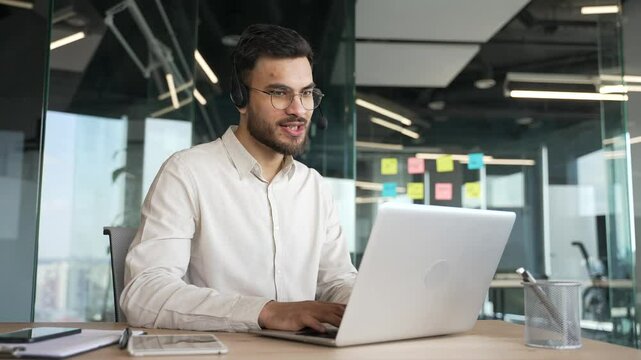 Smiling male corporate operator in headset working in customer support service on helpline telesale talking using laptop in business office. Agent of call center advises client by answering questions