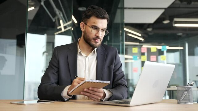 Businessman in a formal suit watching video call conference or training takes notes looking at laptop screen sitting at workplace in business office. Worker on remote certification learning. Skill up