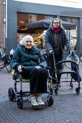 Mature woman walking with a walker next to elderly woman in wheelchair