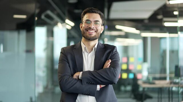 Portrait of smiling handsome businessman in a formal suit standing with crossed arms at workplace in modern business office. Confident successful happy entrepreneur in glasses posing looking at camera