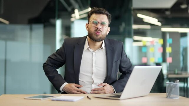 Sick businessman in a formal suit feels stomach pain working on laptop sitting at workplace in business office. Worker has heartburn, gastritis or poisoning. Male suffers from spasms and constipation