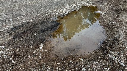 A water-filled pothole on a cracked asphalt road, symbolizing infrastructure decay and weather challenges, with reflections of trees