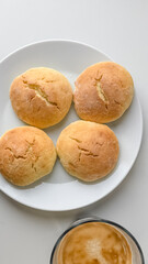 Four freshly baked pandesal rolls on a white plate next to a cup of coffee, celebrating a traditional Filipino breakfast