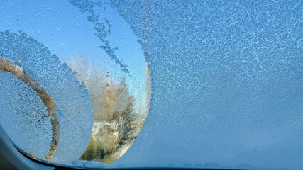A frosted car windshield is partially cleared for driving safety, symbolizing the challenges of...