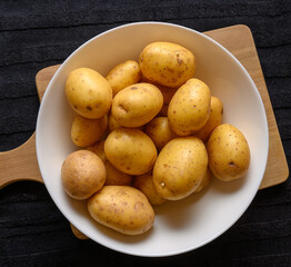 A collection of freshly harvested golden potatoes arranged in a white bowl rests on a wooden cutting board, showcasing their natural textures and earthy tones