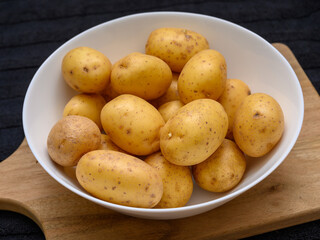 A collection of freshly harvested golden potatoes arranged in a white bowl rests on a wooden cutting board, showcasing their natural textures and earthy tones