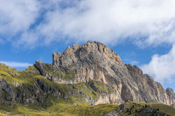 The jagged rocky peaks of Seceda rise sharply above the grassy slopes, creating a dramatic contrast against the bright blue sky and scattered clouds