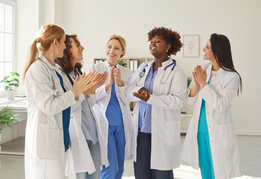 Happy female doctor group celebrating common success, clinic diverse team in white medical professional uniforms in clinic office hall, smiling, praising confident young ambitious women in hospital