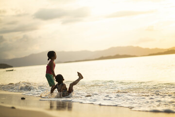 Two children delight in the ocean waves during a dusky sunset, evoking the feelings of warmth, joy,...
