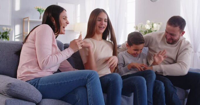 Happy family spending time together on large gray sofa in living room and having fun. Smiling parents tickling and teasing their children, fooling around and enjoying entertainment on weekend at home.