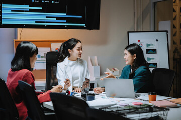 Three asian businesswomen are discussing a project in a meeting room at night, using a laptop and analyzing charts