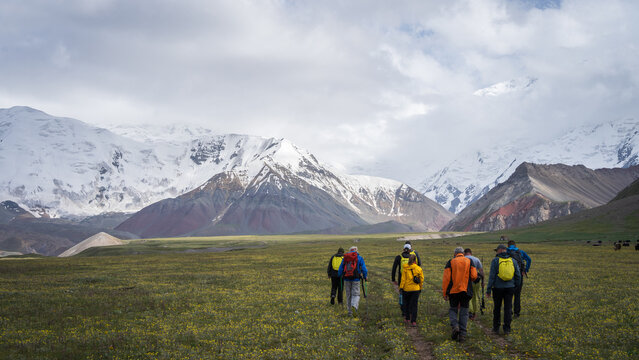 Group of tourists hiking in green alpine valley towards snowy peaks in Pamir Mountains, Kyrgyzstan