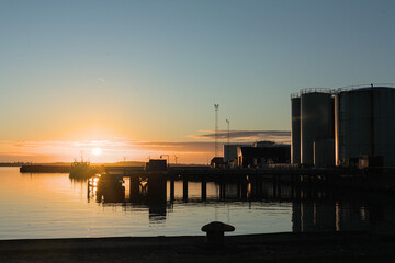 Sunset over industrial harbor with silos and fishing boats reflecting on calm water © Sergei