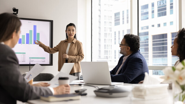 Young Asian businesswoman presenting marketing report on large electronic board to group of colleagues, discussing business plan for project, analyzing sales. Trainer giving corporate seminar