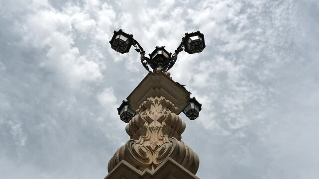 Upward view of Fuente de la Farola, highlighting its intricate stone base and historic four-arm lamp, framed by a bright cloudy sky.