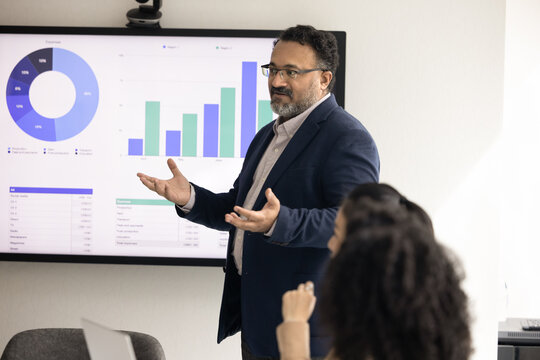 Mature Indian company CEO presenting sales growth analytics to group of employees, colleagues, speaking at electronic board, large wall display in office meeting room, holding presentation