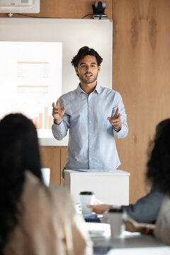 Motivated young Hispanic project manager giving seminar, lecture, presentation at whiteboard, speaking to colleagues at large meeting table, explaining creative business plan. Vertical, shot
