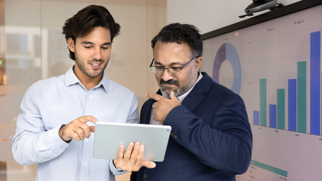 Positive Latin businessman showing project plan on tablet to mature Indian colleague, consulting elder coworkers. Different aged diverse businessmen working with digital gadget together and talking