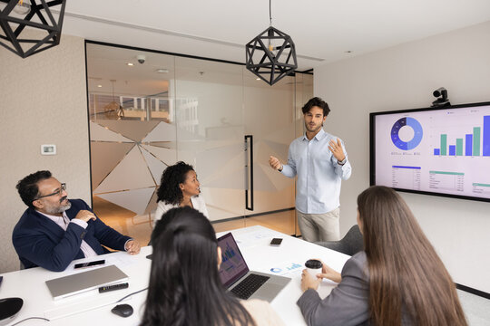 Young Latin professional man holding meeting with diverse team, offering business plan, marketing strategy to colleagues sitting at table, explaining report on large electronic TV screen