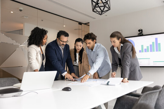 Diverse different aged business team of serious colleagues analyzing financial reports, marketing research results together, bending over meeting table, reviewing paper documents