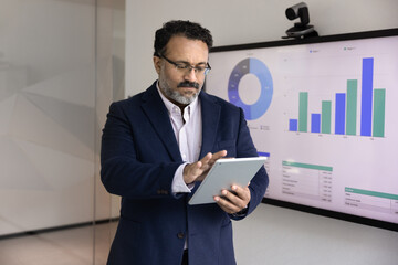 Serious senior Indian business leader man analyzing marketing data on tablet computer, typing on...
